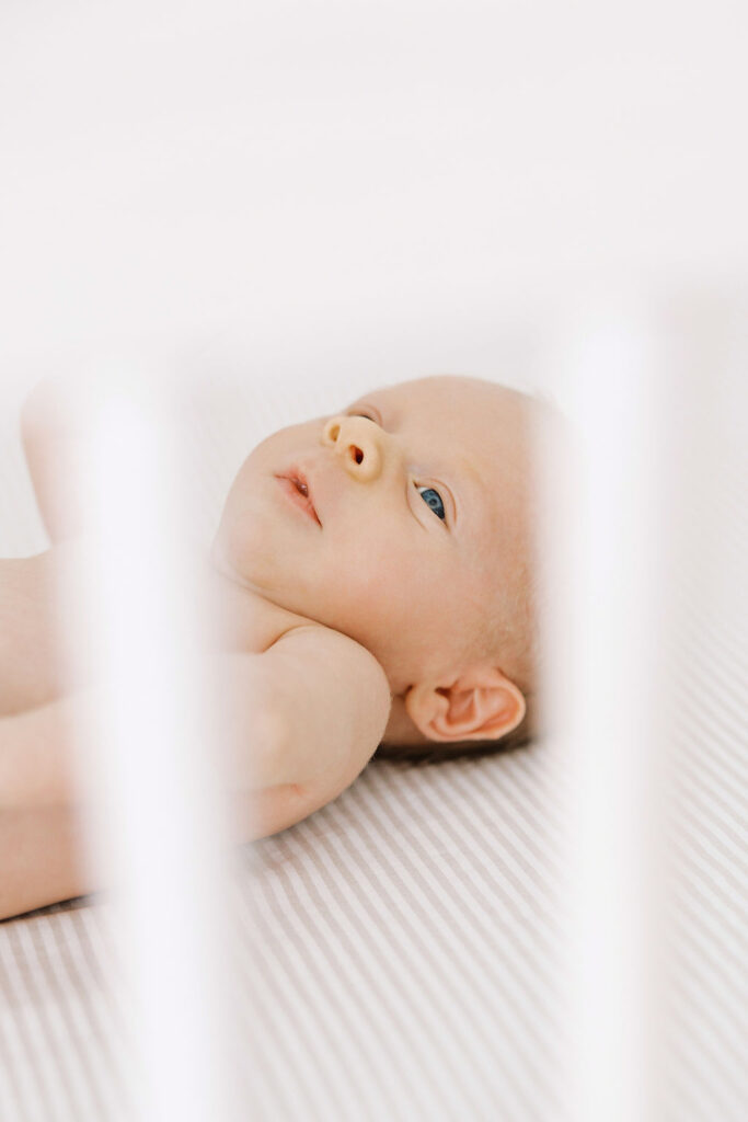 Newborn lying peacefully on a white blanket in his crib and looking upward.
