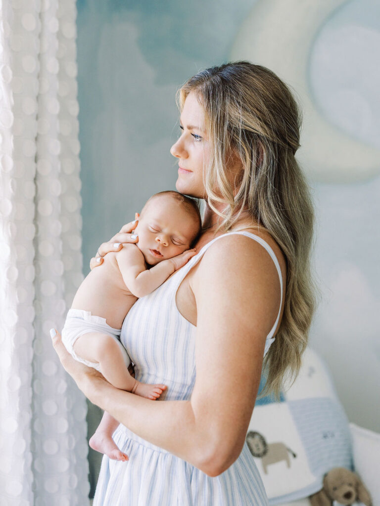 Mother holding her baby near a bright nursery window during an at-home newborn session.