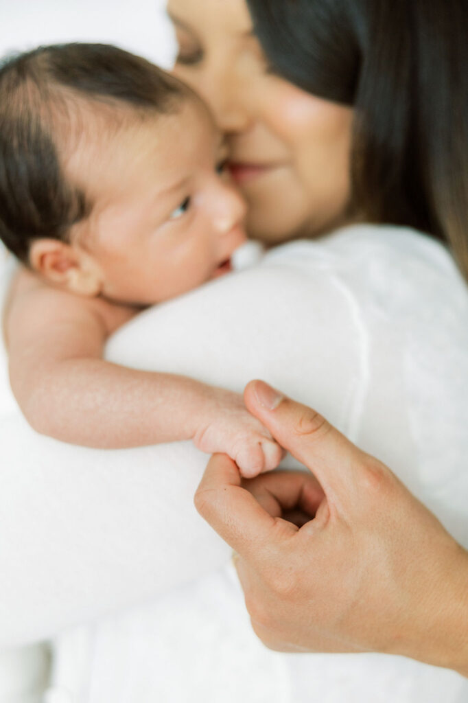 Mom kissing her newborn baby while gently holding the baby’s hand during a studio newborn session.