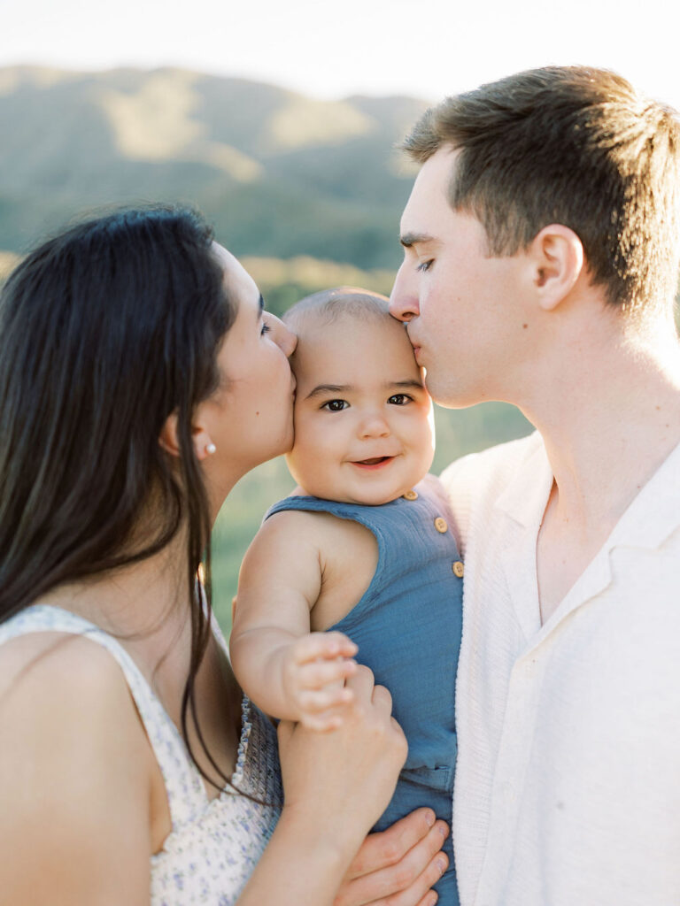 Mom and dad kissing their smiling baby during natural family photos with kids