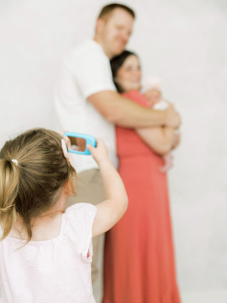 Older sister taking a photo of her parents and newborn baby during a relaxed family newborn session.