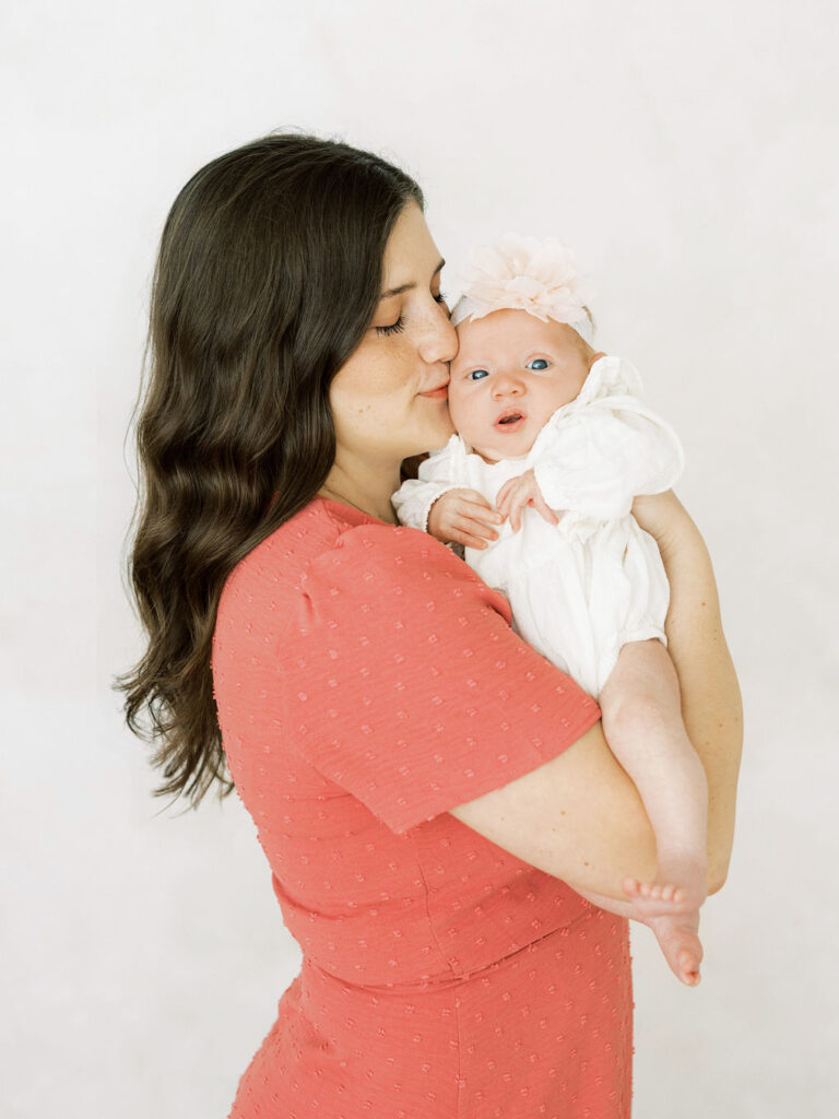 Mom kissing her newborn baby girl while holding her close in a bright, simple setting.