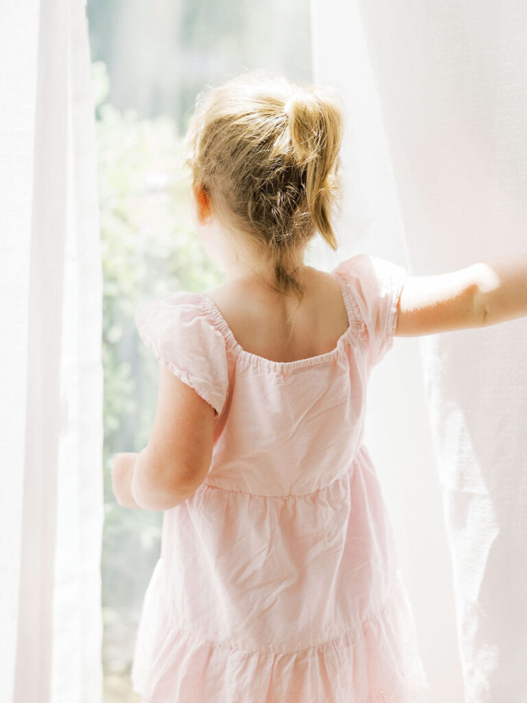 Young girl in a blush pink dress standing by a bright window during a lifestyle newborn session.