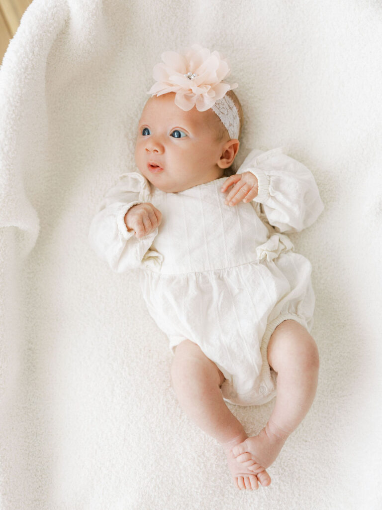 Newborn baby girl lying on a soft neutral blanket in a white outfit and floral headband.