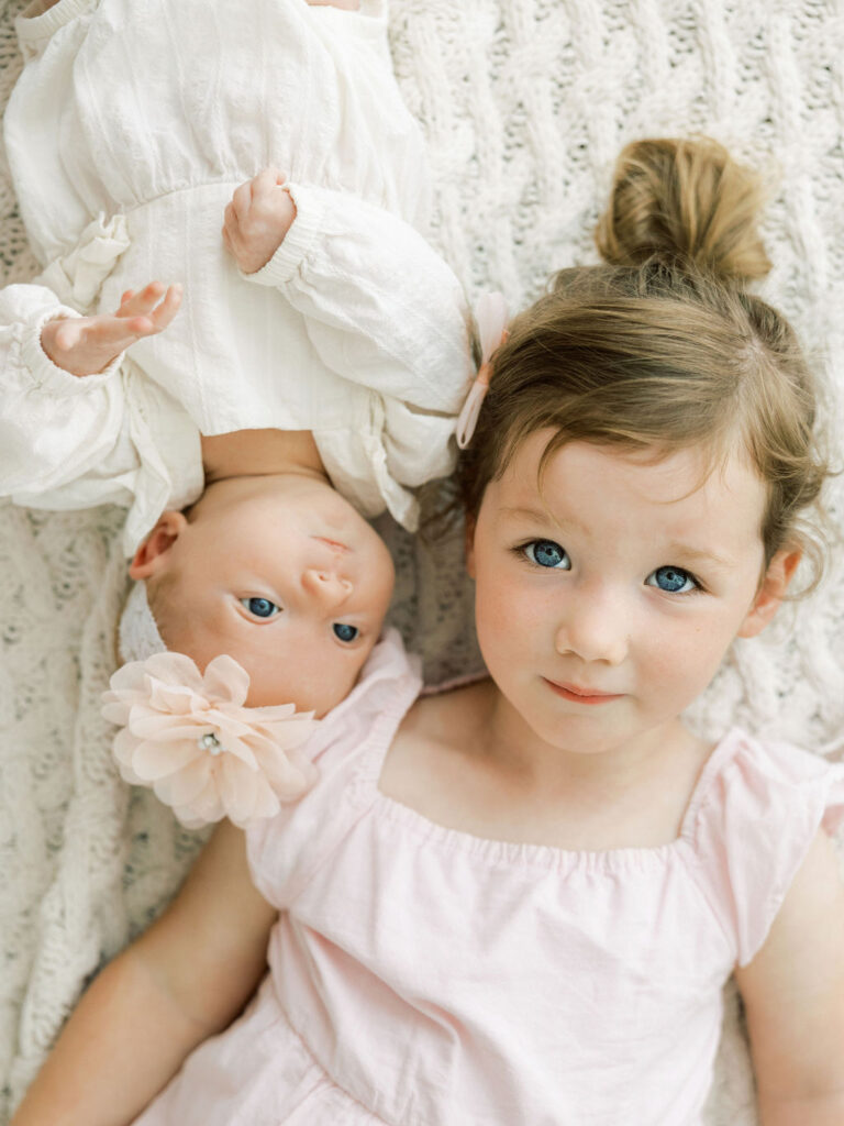 Newborn baby girl lying beside her older sister in soft white and blush outfits.