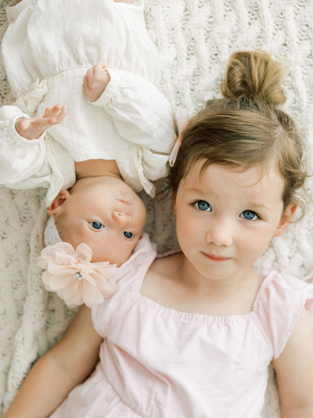 Newborn baby girl lying beside her older sister in soft white and blush outfits.