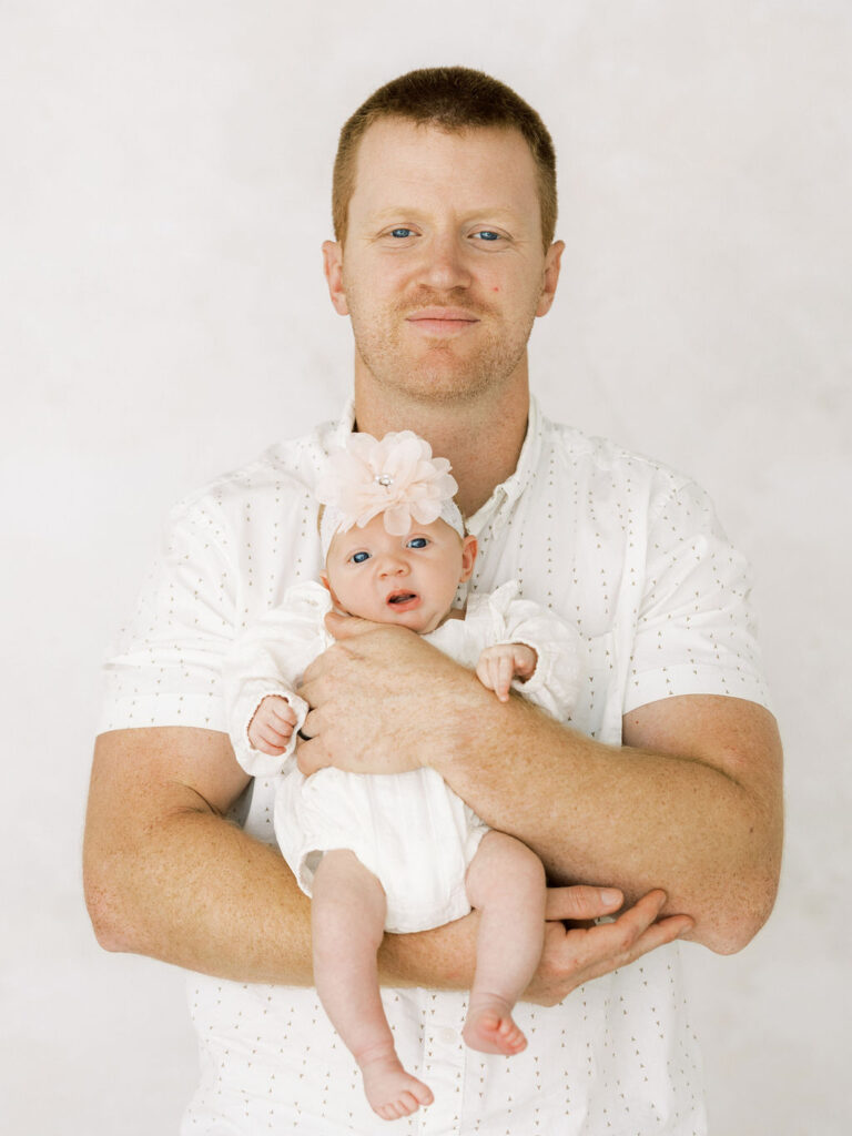 Dad holding his baby girl during newborn photos in Santa Clarita, both dressed in soft white tones for a clean, simple look.