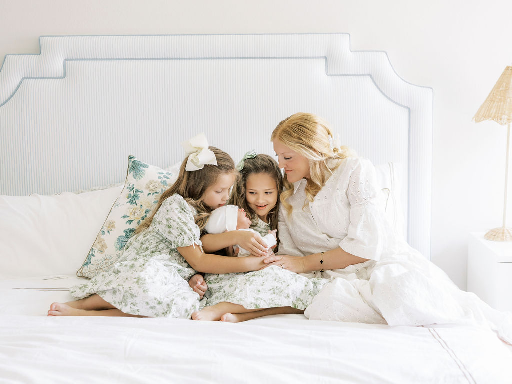 Mom and two older sisters cuddling a newborn baby together on a bed during newborn photos with siblings in Santa Clarita.