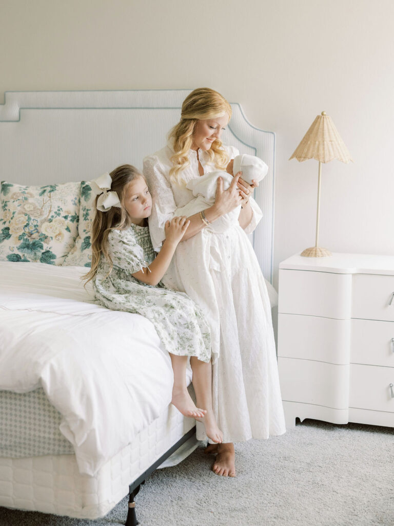 Mom in a white dress holding her newborn baby while an older sister stays close during a natural newborn photos with siblings session.