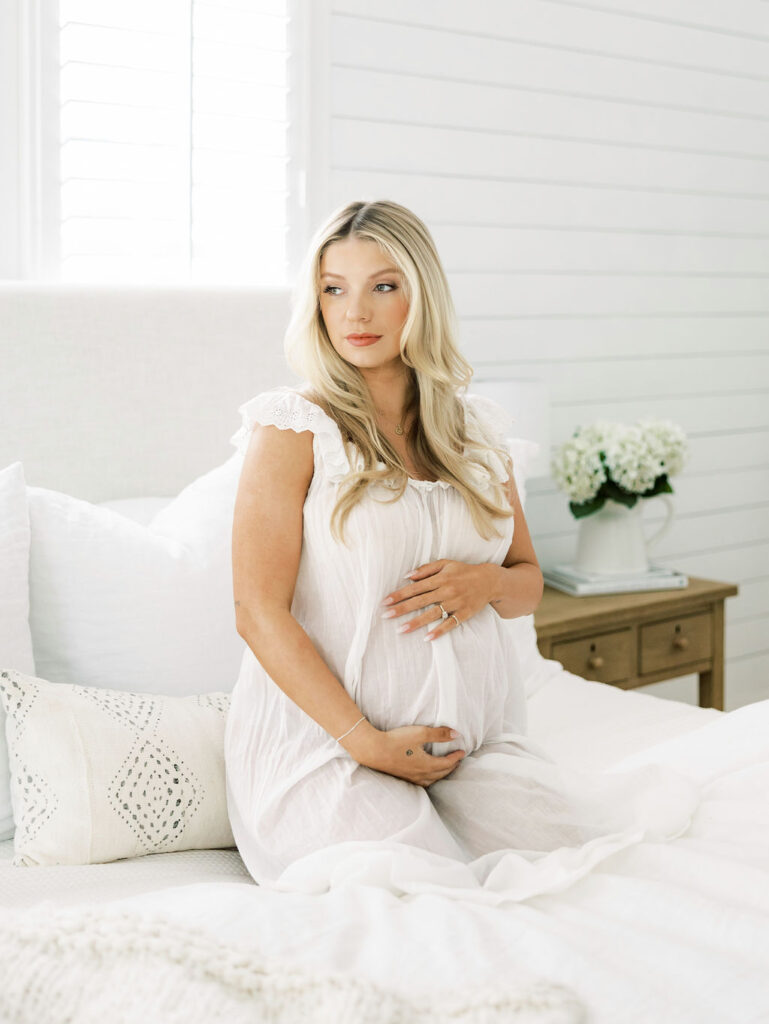 Pregnant woman standing in a light filled room in a simple neutral look for her maternity photography session