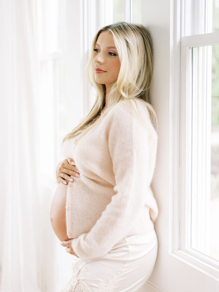 Pregnant woman standing beside a window in a soft neutral outfit during a Santa Clarita maternity photo session