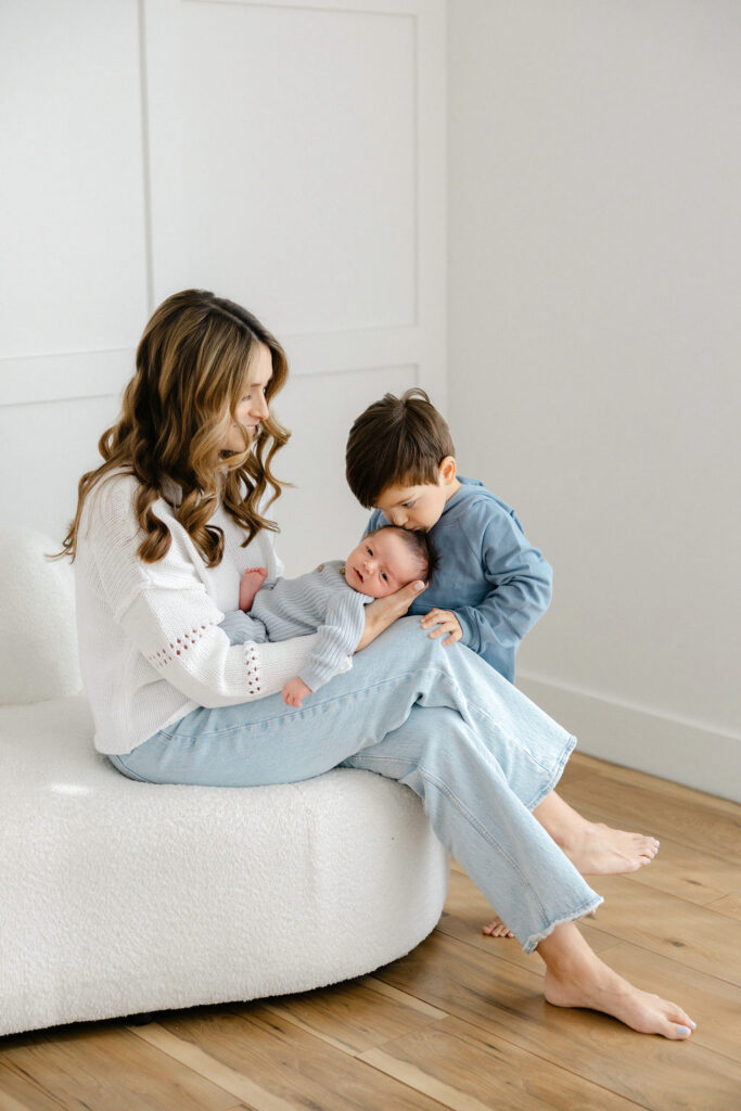 Mom holding her newborn baby while an older sibling gives the baby a gentle kiss during a studio newborn session.