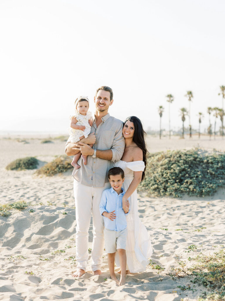 Family of four standing together on the beach in coordinated white, light blue, and neutral outfits for a polished family photo look.