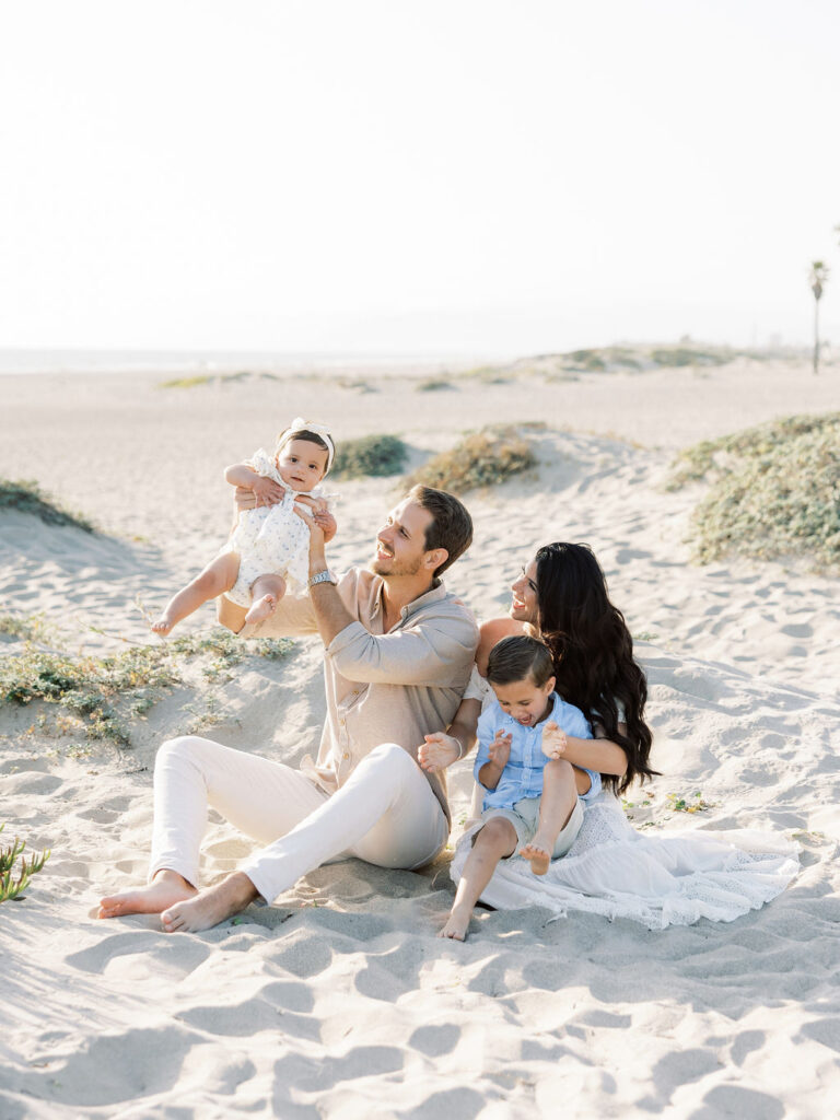 Family of four relaxing together on the beach in coordinated white, light blue, and neutral outfits for a soft family photo look.