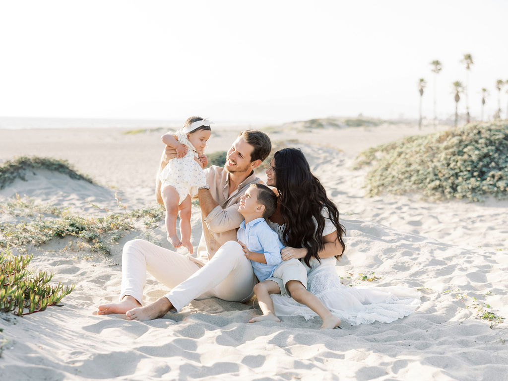 Family of four sitting together on the beach in coordinated white, light blue, and neutral outfits with soft textures.