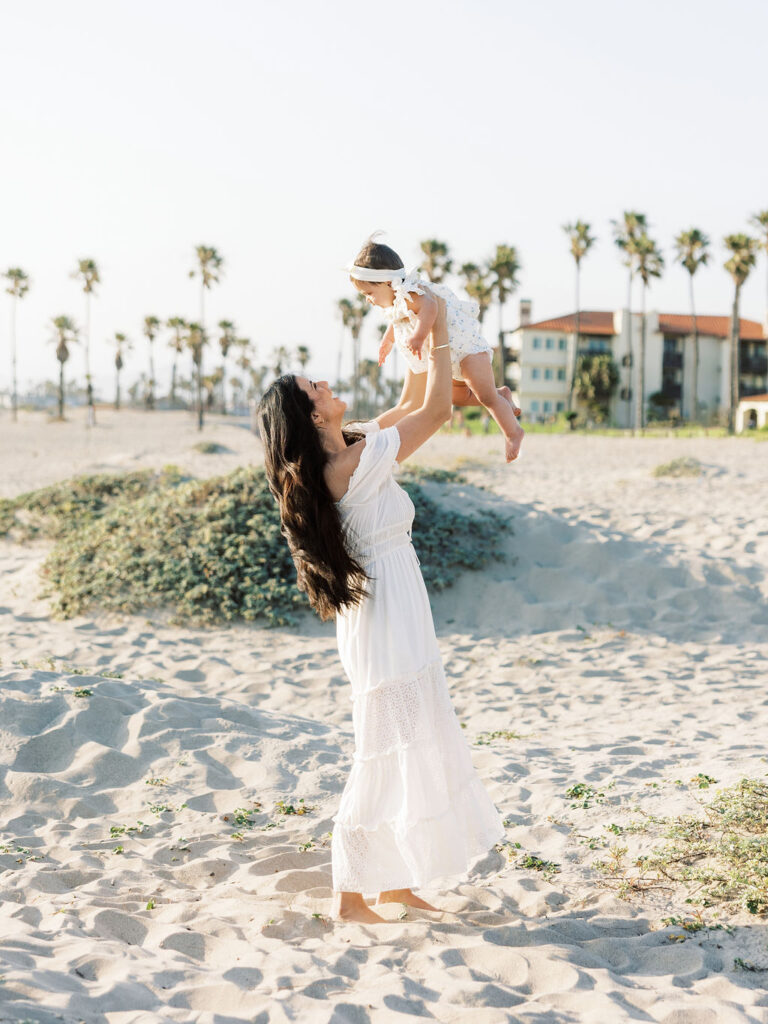 Mom lifting her baby girl on the beach, with soft white outfits and delicate textures creating a simple coordinated look.