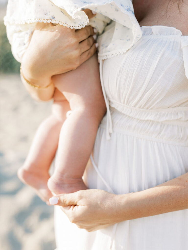 Close detail of a baby’s legs in a soft floral outfit against mom’s white dress, highlighting light textures for family photo styling.