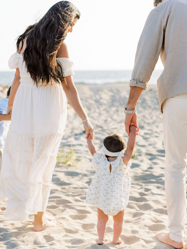Baby girl walking between her parents on the beach in coordinated white and neutral family photo outfits.