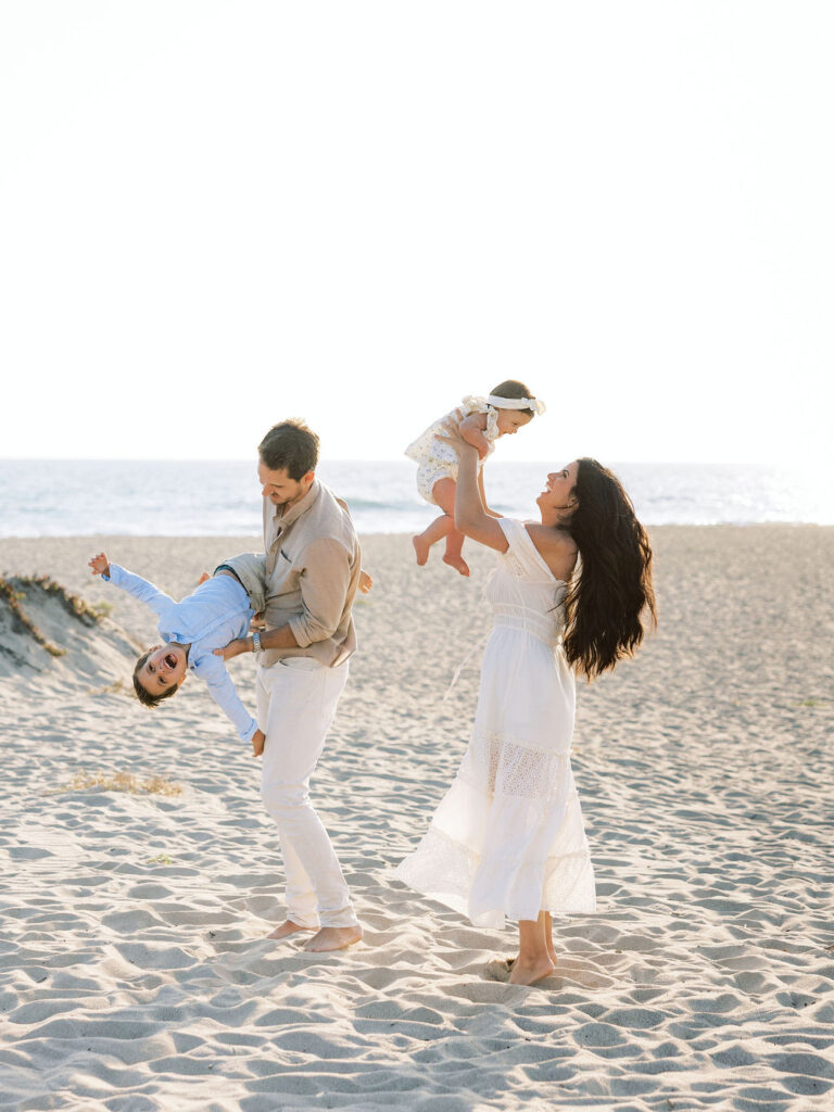 Family playing together on the beach in coordinated white, light blue, and neutral outfits.