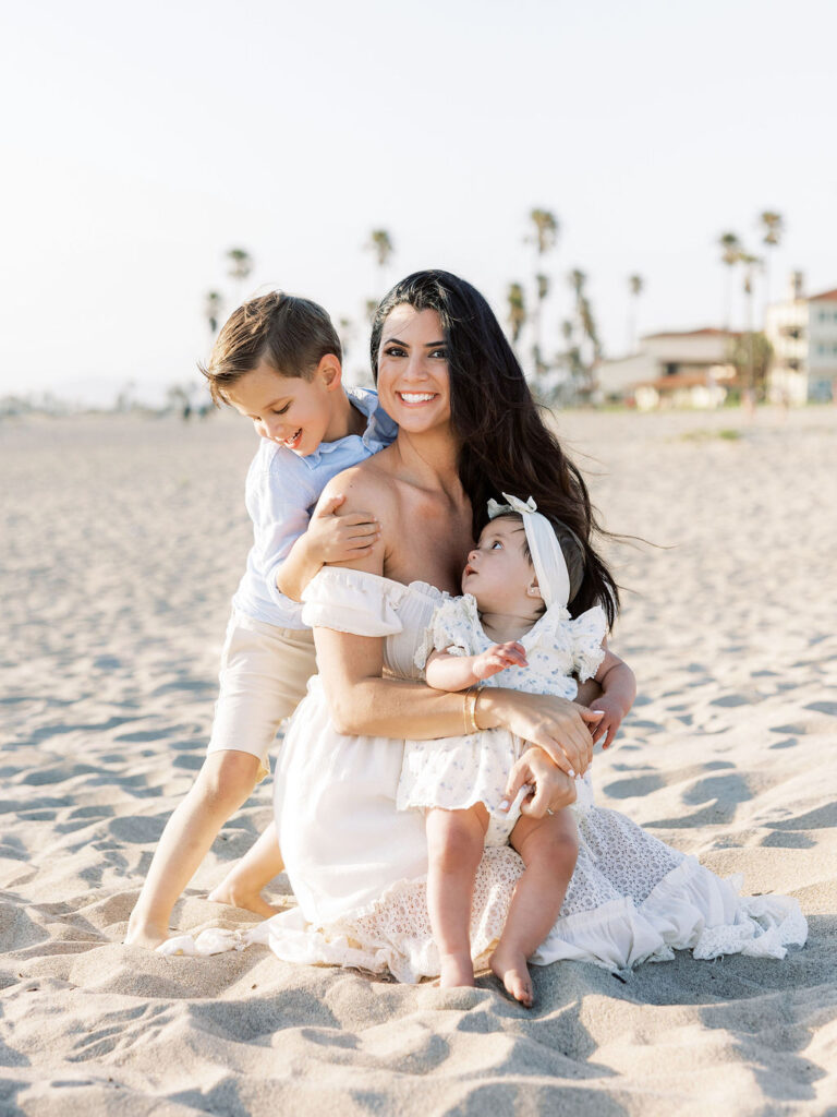 Mom sitting on Oxnard Beach with her son and baby girl in coordinated white, light blue, and neutral outfits.