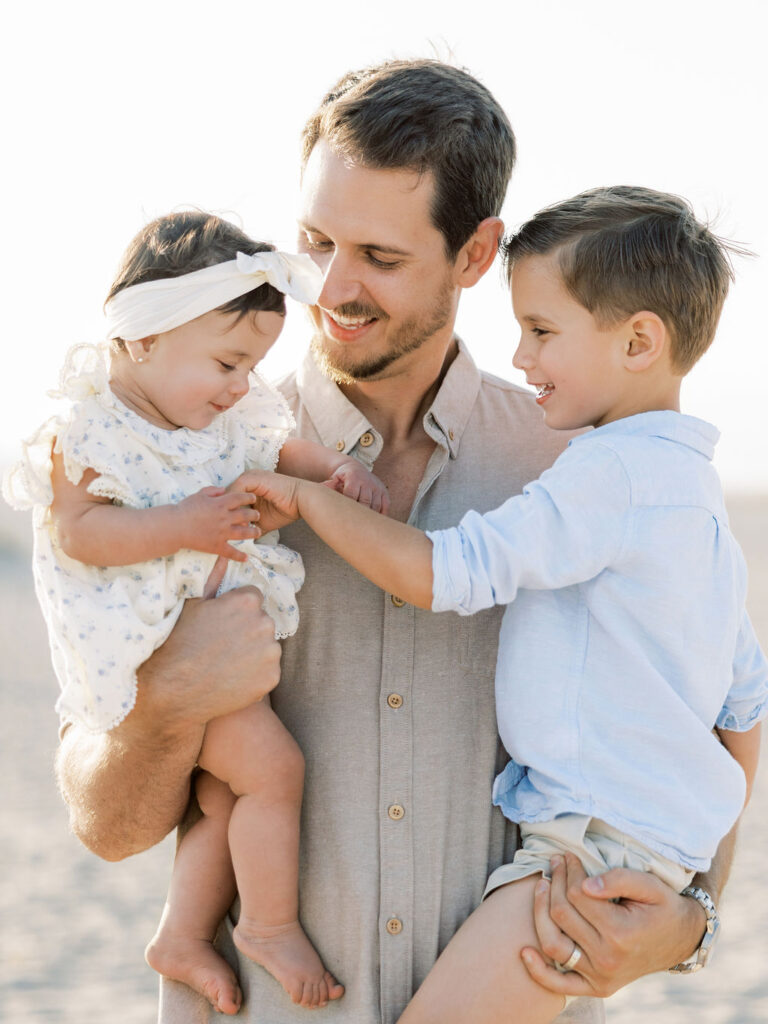 Dad holding his baby girl and older son on Oxnard Beach in coordinated neutral, white, and light blue outfits.