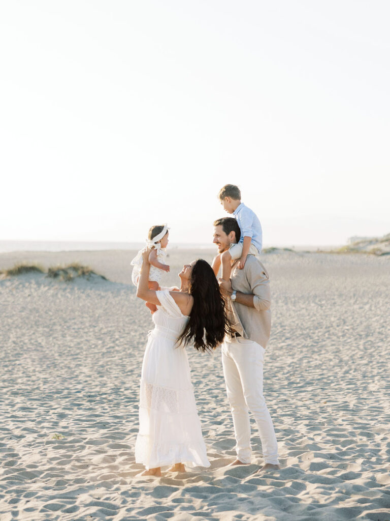 Family of four playing together on Oxnard Beach in coordinated white, light blue, and neutral outfits.