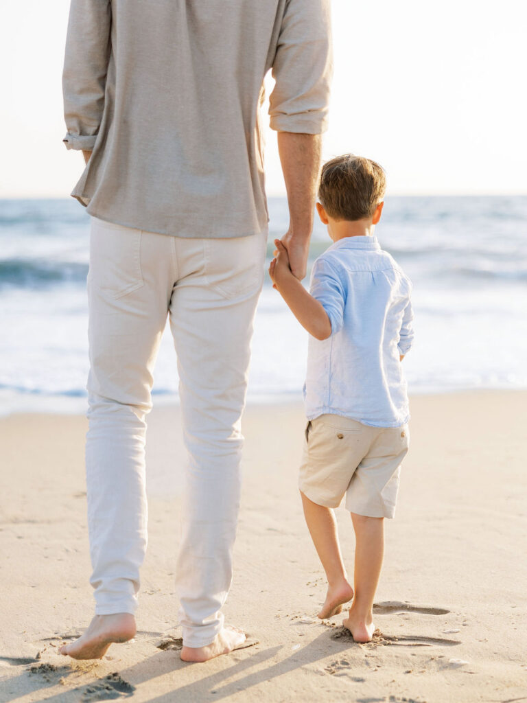 Dad and son holding hands on Oxnard Beach in coordinated neutral and light blue outfits.