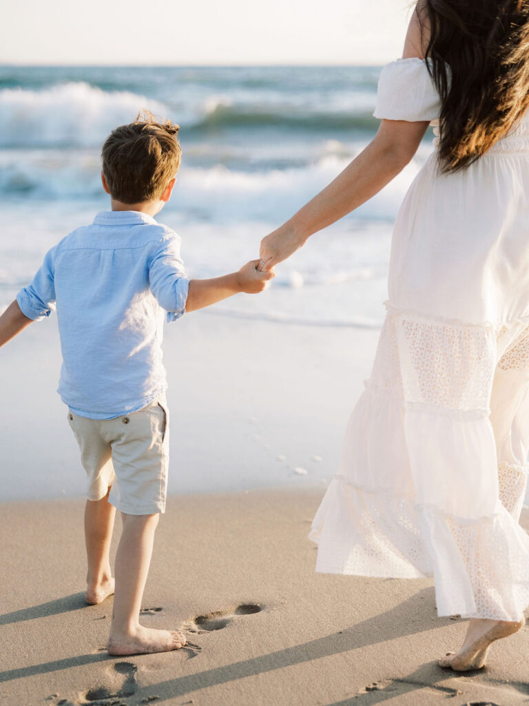 Mom and son holding hands as they walk toward the ocean at Oxnard Beach in coordinated white and light blue outfits.
