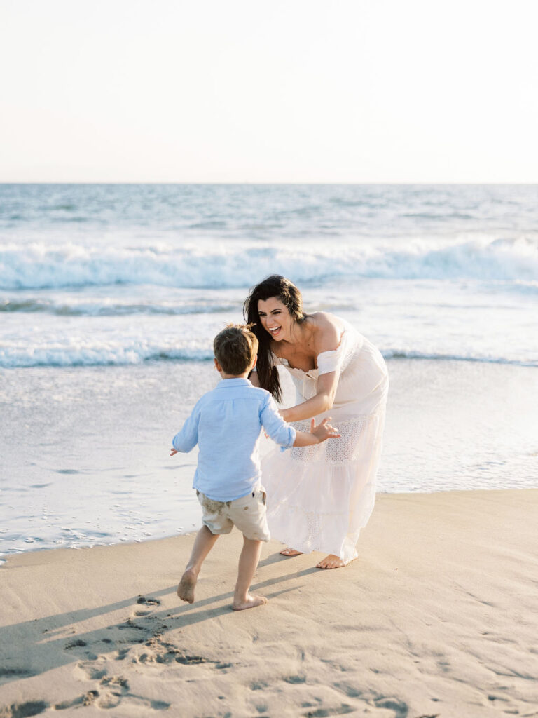 Mom smiling as her son runs toward her on Oxnard Beach, wearing coordinated white and light blue outfits.