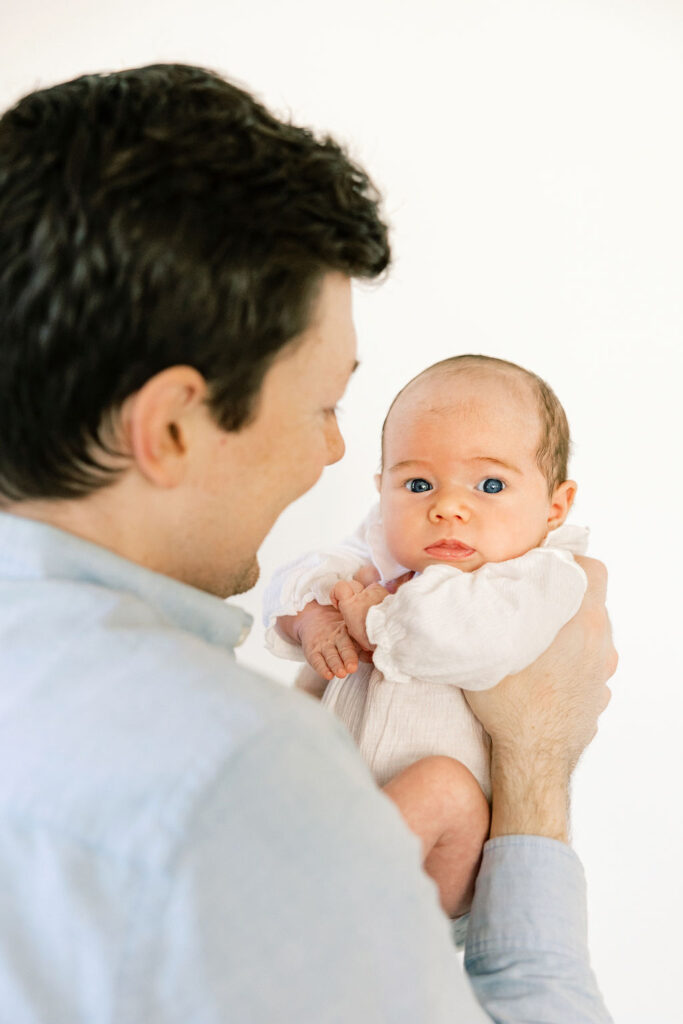 Dad holding his newborn in a soft white outfit, showing simple neutral newborn photo styling