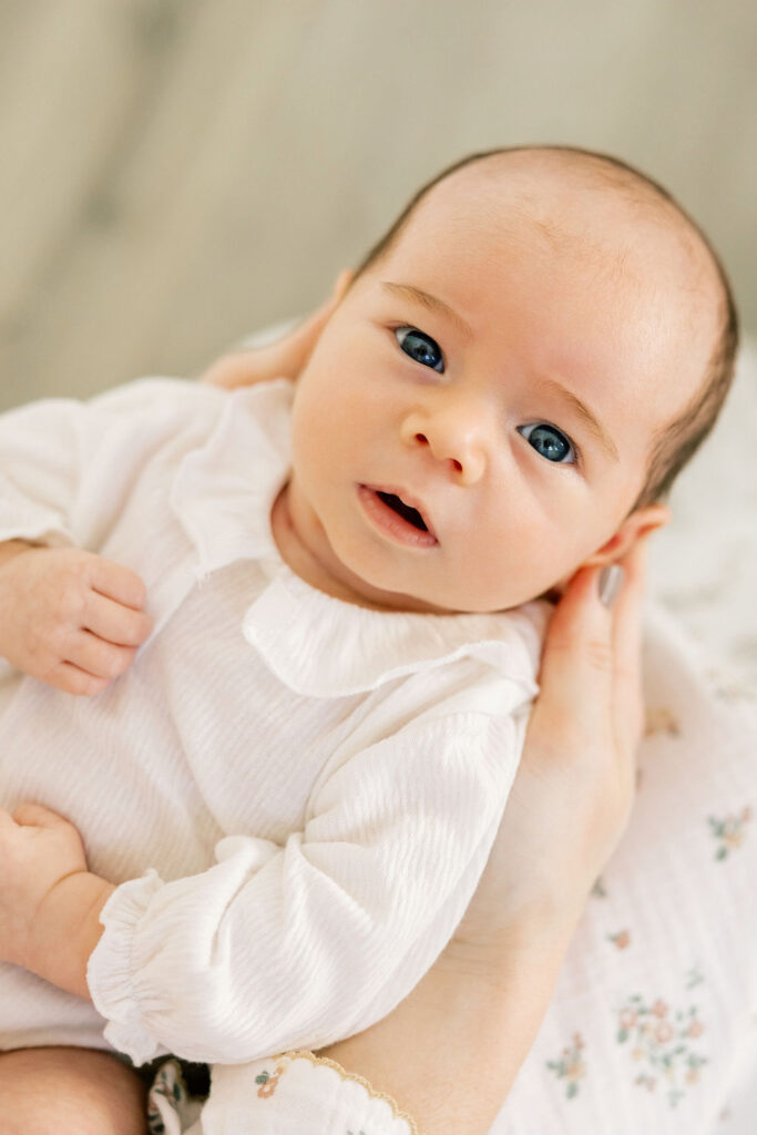 Newborn baby in a soft white outfit held in mom’s arms during a simple, timeless newborn photo session