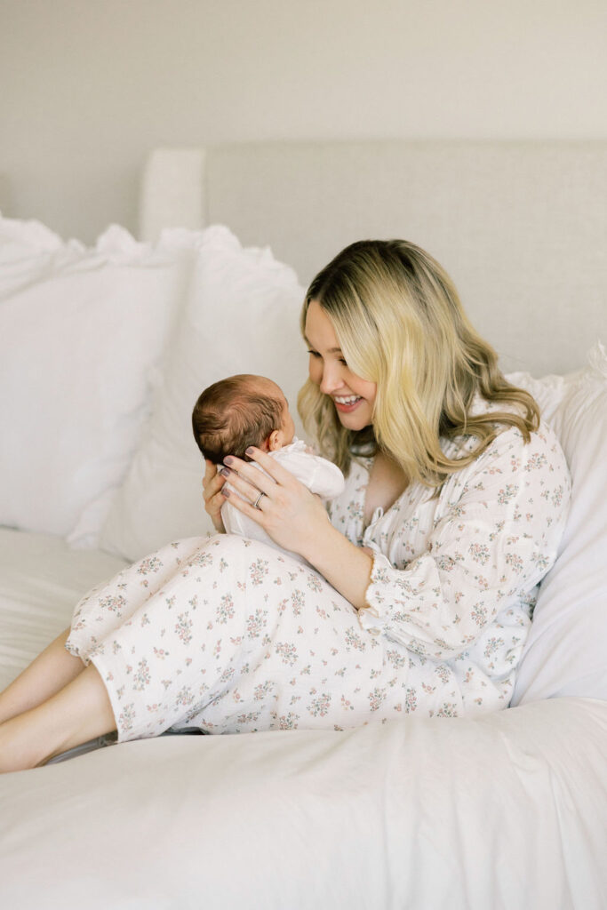 Mom cuddling her newborn on a white bed while wearing a soft floral dress, a timeless outfit choice for in home newborn photos in Santa Clarita
