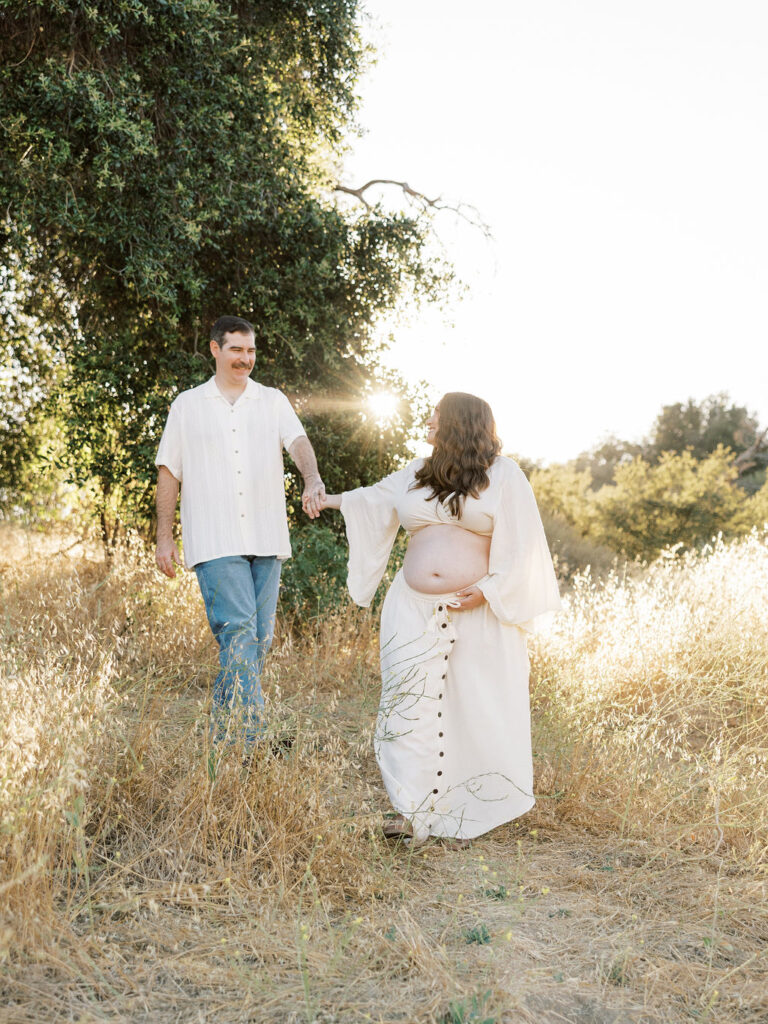 When to Take Maternity Photos with an expecting couple walking hand in hand through a golden Santa Clarita field