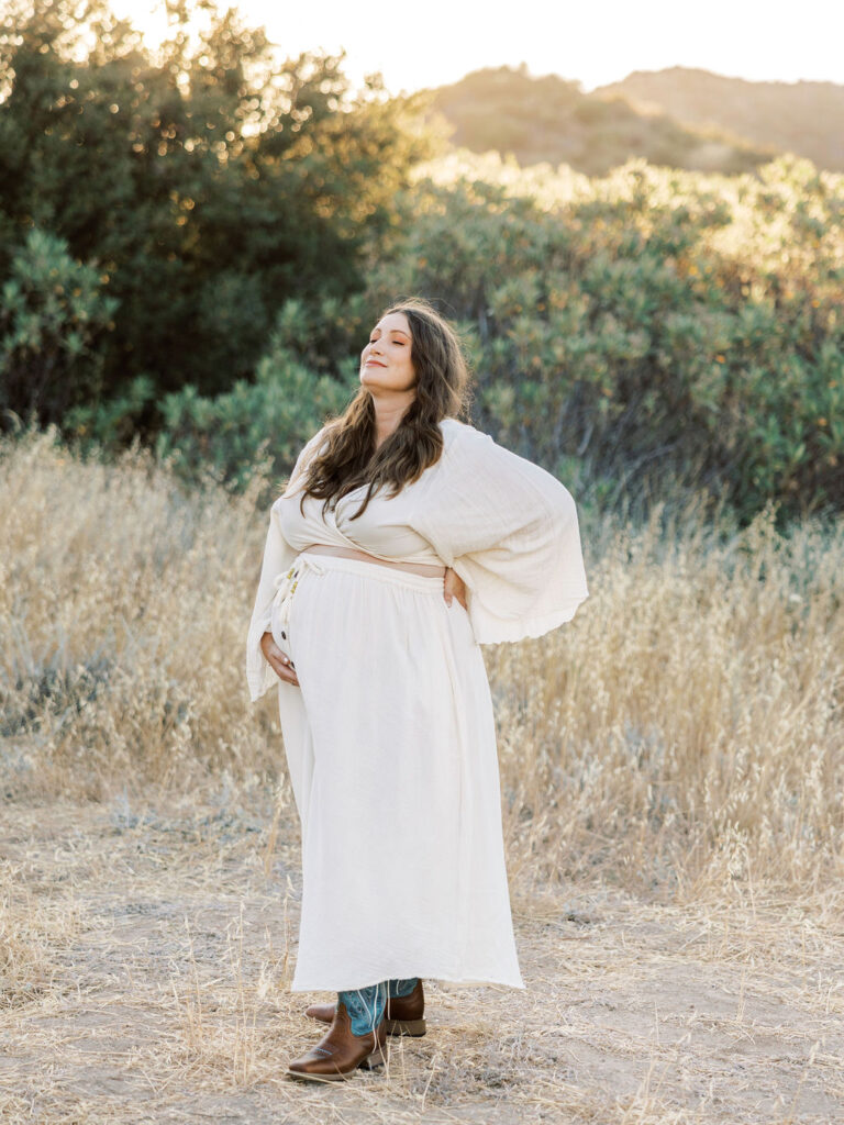 Pregnant woman in a flowing white dress standing in a golden field during a Santa Clarita maternity photo session