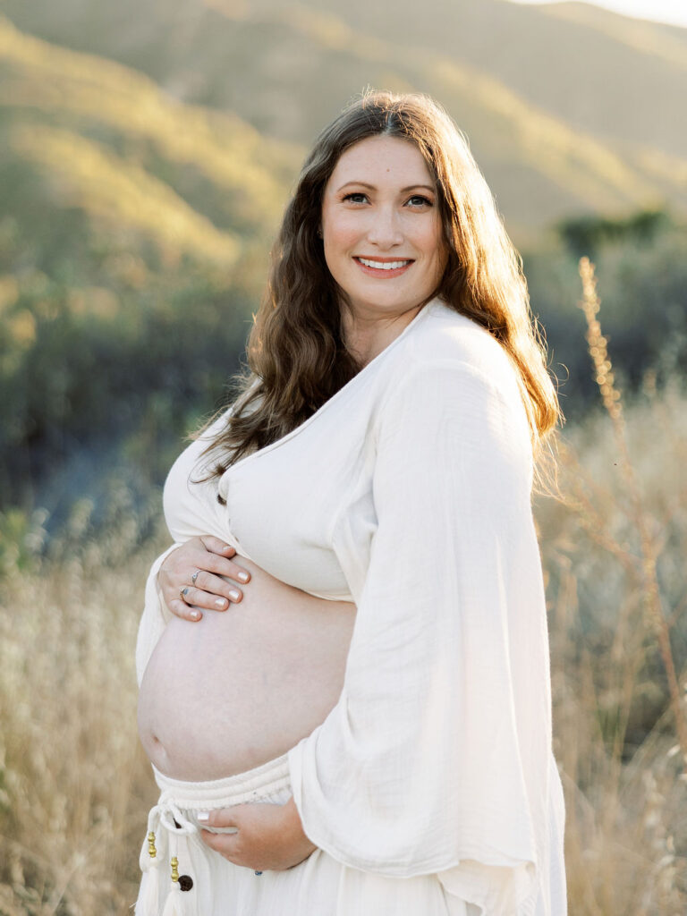 Pregnant woman smiling in a white dress during her maternity portraits